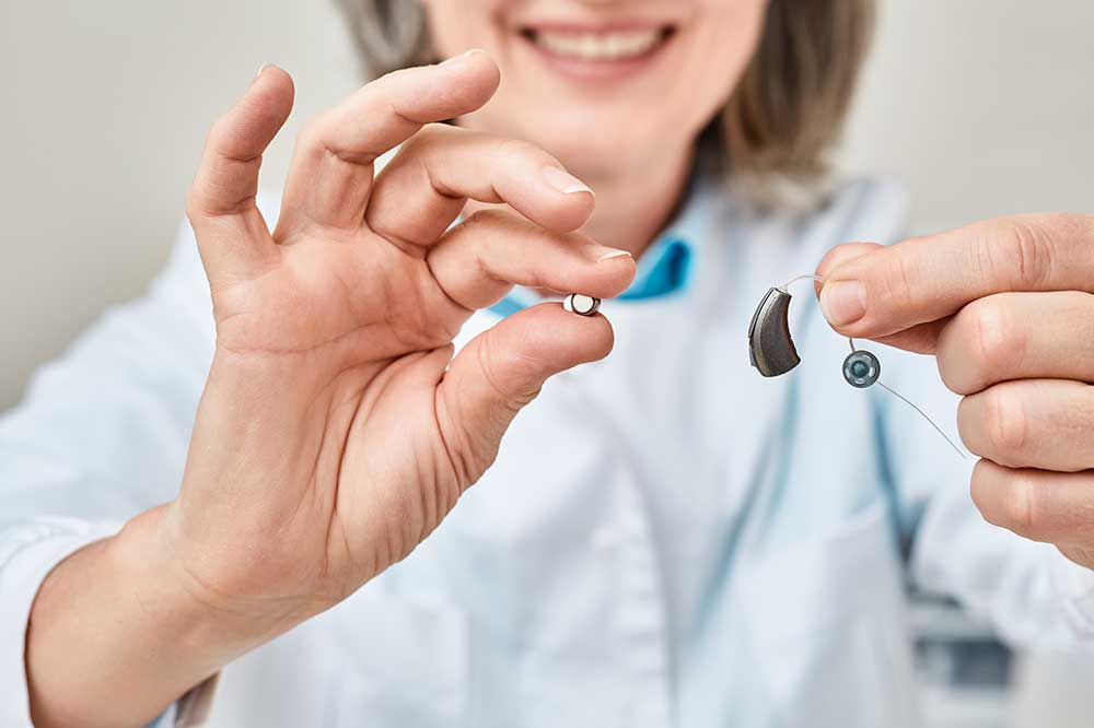Smiling hearing care professional holding up a battery and a receiver-in-canal hearing aid, for battery tips from Gomer Hearing Center in Richardson, TX