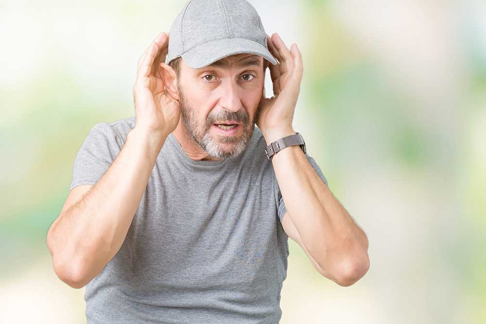 Man in a gray cap with hands cupped around his ears, straining as if one side is quieter, a concern Gomer Hearing Center checks in Richardson, TX
